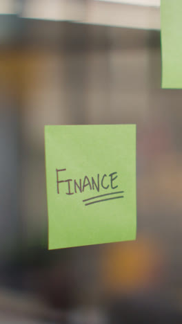 Vertical-Video-Close-Up-Of-Woman-Putting-Sticky-Note-With-Finance-Written-On-It-Onto-Transparent-Screen-In-Office